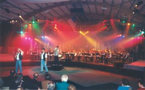 Photo du concert spectacle de 1997 présentant la scène avec 2 chanteurs et en arrière plan l'harmonie elle-même